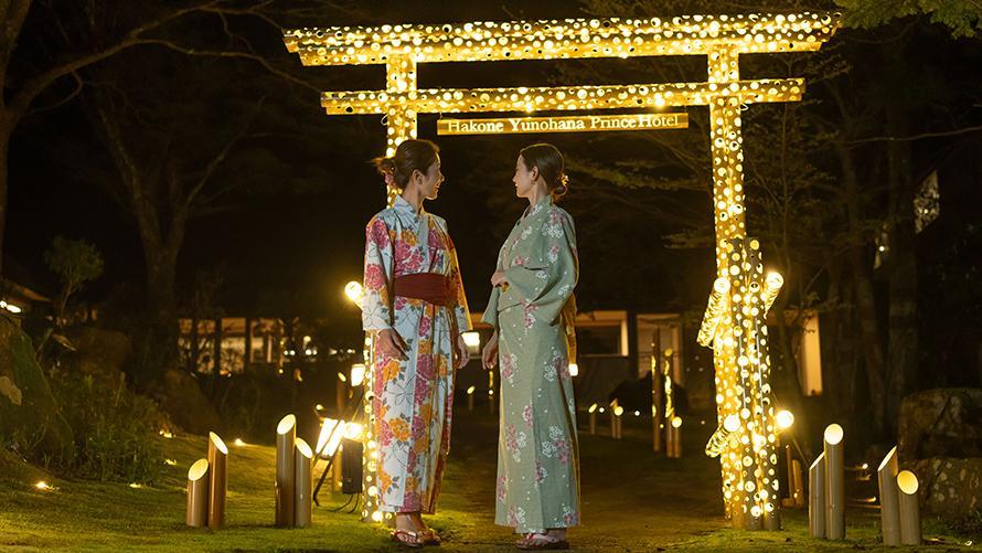 浴衣を着た2人の女性が、光で装飾された大きな鳥居の前で立ち話をしています。  周囲には柔らかな光を放つ照明が配置され、静かでロマンチックな夜の雰囲気を演出しています。